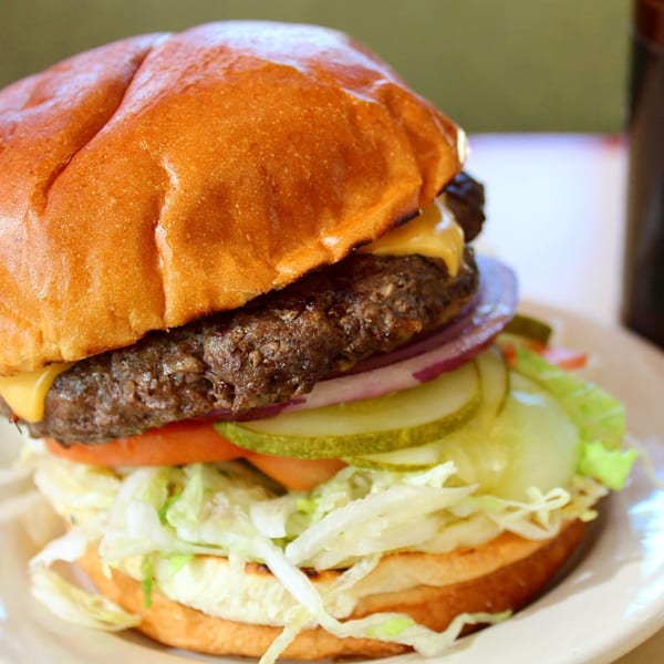 A Hub's Restaurant cheeseburger with lettuce, tomato, onion, and pickles on a white plate.