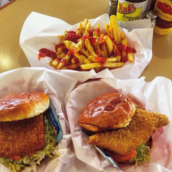 Two Hub's fish sandwiches with lettuce and tomato, alongside a basket of french fries with ketchup.