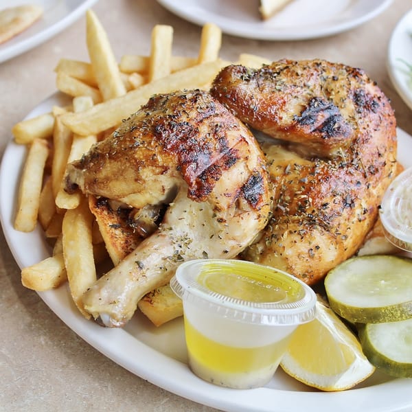 Hub's Restaurant's Greek chicken with crispy fries, lemon, pickles, and dressing on a white plate.