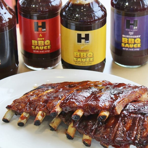 Glazed BBQ ribs on a white plate with three bottles of Hub's BBQ sauce in the background.